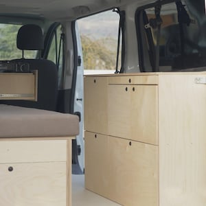 Può includere: Vista interna di un furgone bianco con mobili in legno personalizzati. I mobili includono cassetti e vani portaoggetti. Sono visibili anche una piattaforma letto e un piccolo piano di lavoro. L'interno del furgone è progettato per il campeggio o i viaggi.