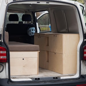 Può includere: Vista interna di un furgone bianco con mobili in legno personalizzati. L'arredamento comprende una piattaforma letto con cuscino marrone, cassetti e armadi. Un casco blu è appeso al poggiatesta. Il portellone posteriore del furgone è aperto.