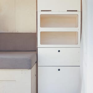 Op de afbeelding: Een ingebouwde witte houten kast met twee open planken en twee laden. De kast heeft een natuurhouten frame en zwarte knoppen. Links is een grijze bank met kussens.