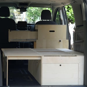 Può includere: Un kit di conversione per camper in compensato chiaro installato nel retro di un veicolo. L'unità presenta una rete a doghe piatta, un grande cassetto estraibile con guide metalliche e un mobiletto con una manopola di colore nero.