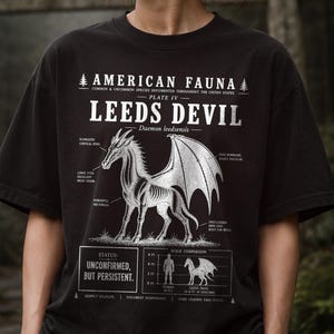 May include: Black t-shirt featuring a detailed illustration of the Leeds Devil, a mythical creature, with anatomical labels and the text "AMERICAN FAUNA." The design includes a scale comparison and the phrase "UNCONFIRMED, BUT PERSISTENT."