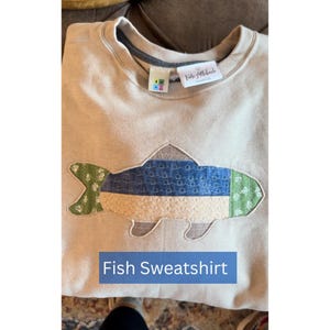 May include: A beige sweatshirt with a fish applique. The fish design features blue, green, and beige patterned fabric. The text "Fish Sweatshirt" is in a blue rectangle at the bottom of the image. The sweatshirt has a crew neck.