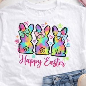 May include: White t-shirt featuring three colorful tie-dye bunny silhouettes with bows, flowers, and hearts. The words "Happy Easter" are written in pink script below the bunnies. The shirt is paired with blue jeans and sneakers.
