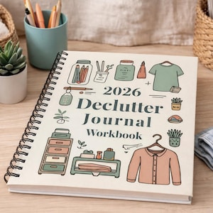May include: A spiral-bound 2026 Declutter Journal Workbook with illustrations of organizing tools and clothing. The cover is cream-colored with black text and drawings in green, peach, and brown. A succulent plant in a white pot is visible.