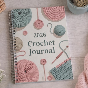 Puede incluir: Un diario de ganchillo 2026 con espiral y cubierta crema, con ilustraciones de ovillos de lana y elementos de ganchillo en rosa, verde y beige. El diario está rodeado de hilo, una taza y una manta de punto rosa.