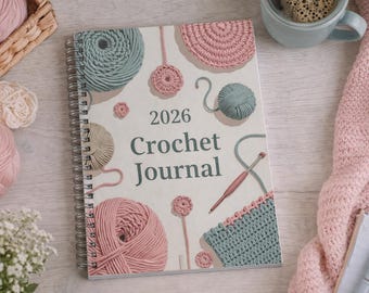 May include: A spiral-bound 2026 Crochet Journal with a cream-colored cover featuring illustrations of yarn balls and crochet elements in pink, green, and beige. The journal is surrounded by yarn, a cup, and a pink knitted blanket.