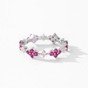 May include: A silver-toned band ring featuring a repeating pattern of four-petaled flower motifs. Each flower is composed of four round, deep pink stones with a single, smaller, round, light pink stone set between each floral cluster.