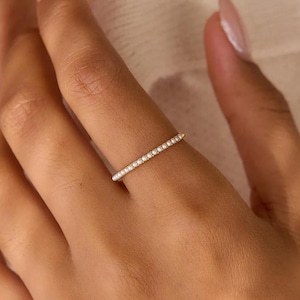 Pode incluir: Um anel de banda fina em ouro com uma fila de pequenas pérolas brancas redondas colocadas juntas ao longo da metade superior da banda. O anel é usado num dedo, destacando o design delicado e o contraste entre o ouro e as pérolas.