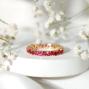 May include: A gold eternity band featuring a continuous row of vibrant, round-cut red rubies set in a shared-prong setting. The ring is positioned horizontally, showcasing the uniform size and deep crimson color of the gemstones.