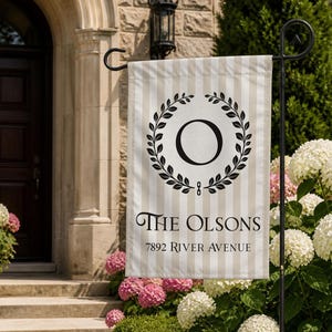 May include: A vertical garden flag featuring a beige and white striped background. The center displays a black circular laurel wreath enclosing the letter O. Below the wreath, the text reads THE OLSONS and 7892 RIVER AVENUE in black.