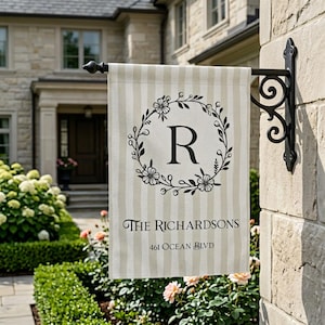 May include: A beige and white striped house flag with a black floral wreath and the letter "R". The flag also displays the text "THE RICHARDSONS" and "461 OCEAN BLVD". The flag is mounted on a black metal bracket.