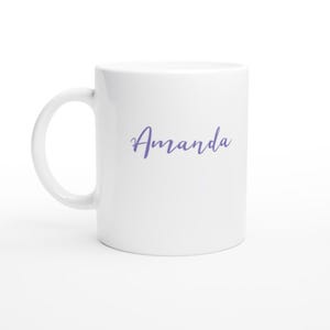 May include: White ceramic mug with a curved handle. The word "Amanda" is written in a cursive, light purple font across the front of the mug. The mug is clean and simple.