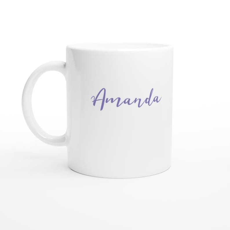 May include: White ceramic mug with a curved handle. The word "Amanda" is written in a cursive, light purple font across the front of the mug. The mug is clean and simple.
