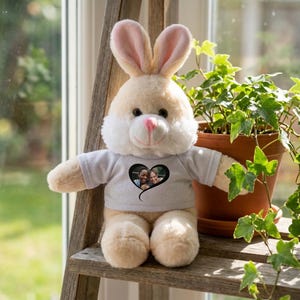 Puede incluir: Un conejo de peluche con pelaje color crema, orejas rosas y nariz rosa. El conejo lleva una camiseta gris claro con un gráfico en forma de corazón negro que contiene una foto. El conejo está sentado sobre una superficie de madera.