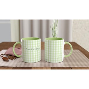 May include: Two white ceramic mugs with a green gingham pattern and green handles. The name "Samantha" is written in cursive on one mug. The mugs are on a checkered placemat.