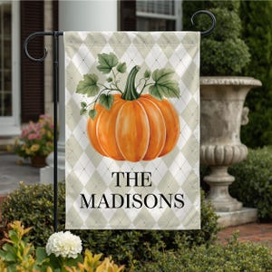 May include: A rectangular garden flag featuring a light beige and white argyle pattern background. Centered is a large orange pumpkin with green leaves and a stem. The black text reads THE MADISONS in a serif font below the pumpkin.