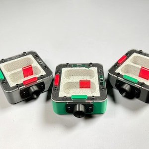 May include: Three square ant farms in gray and black, with a green or gray base. Each has a clear top, a light-coloured interior, and red and green accents. A black tube extends from the front of each farm.