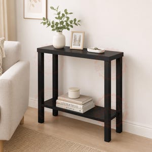 May include: A black rectangular console table with four straight legs and a lower shelf. The top surface holds a white ribbed vase with green foliage, a small framed picture, and a white tray. The lower shelf holds two stacked books.