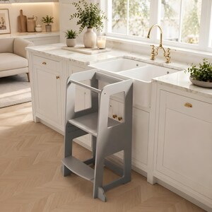 May include: A grey and white wooden kitchen step stool with two steps and a safety bar. The step stool is designed to help children reach work surfaces. The background shows a white kitchen with a marble worktop and a double sink.