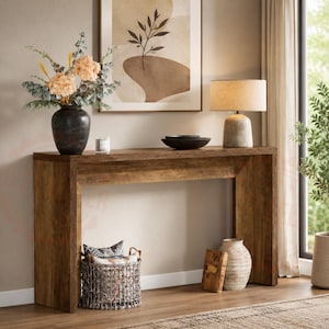May include: A rectangular wooden console table with a natural grain finish. The table surface holds a dark vase with flowers, a small candle, and a black bowl. A woven basket with patterned pillows and a ceramic vase sit on the floor below.