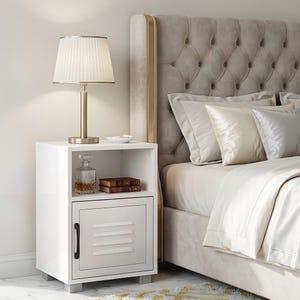 May include: A white bedside cabinet with a metal door and a shelf. A lamp with a pleated shade sits on top, along with a small bowl. A decanter and books are visible on the shelf. The bed has a tufted headboard and pillows.