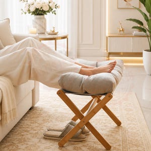 May include: A light grey footstool with a padded top and a folding wooden frame. The footstool is positioned in front of a sofa, with a person's feet resting on it. A pair of slippers sits on the floor in front of the stool.