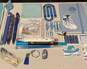 以下が含まれることがあります： 青色の文房具の詰め合わせ。ペン、ペンケース、ノート、ステッカーなど。ペンケースは白のドット柄のライトブルー。ノートには木の模様とクラゲのステッカーが付いています。クリップやキーホルダーもあります。