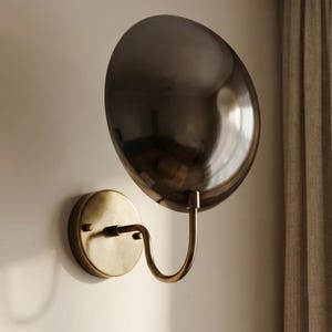 May include: A wall-mounted lamp featuring a circular, dark metallic reflective shade attached to a curved arm. The base is a round, brushed brass-toned plate that secures the fixture to the wall, connecting to a slender, J-shaped metal arm.