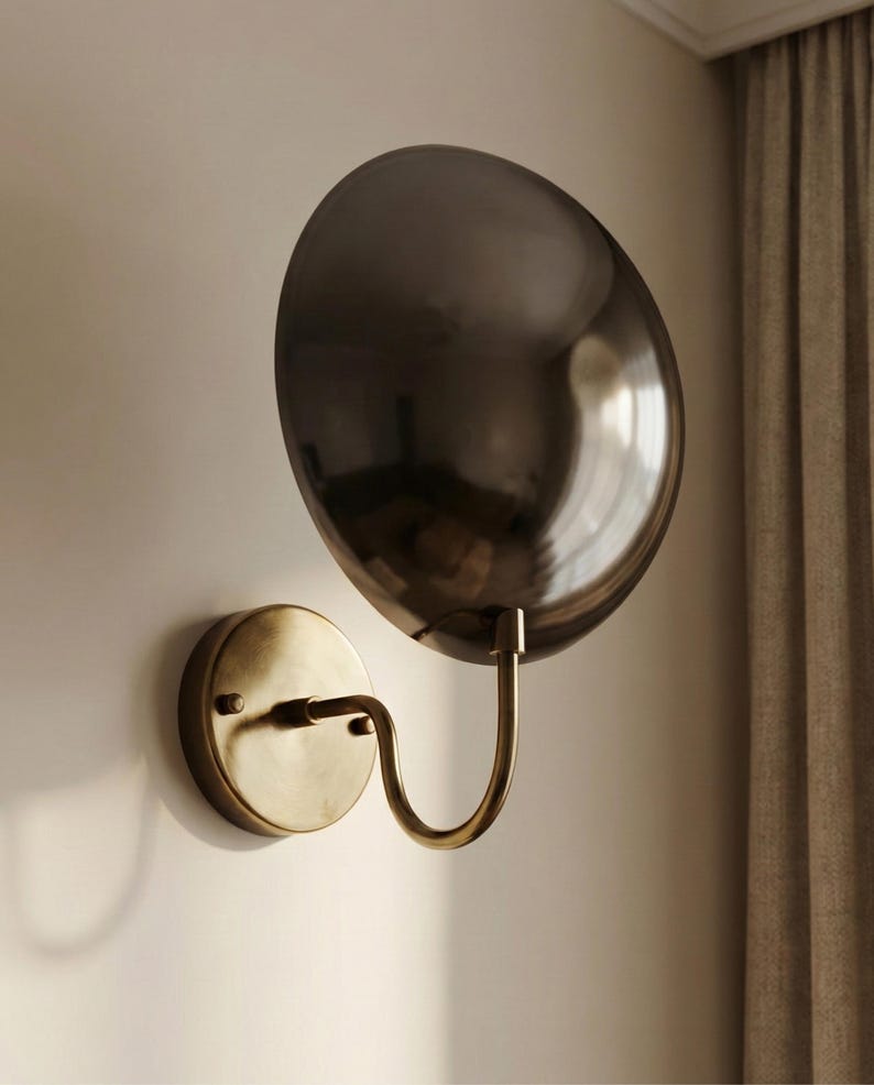 May include: A wall-mounted lamp featuring a circular, dark metallic reflective shade attached to a curved arm. The base is a round, brushed brass-toned plate that secures the fixture to the wall, connecting to a slender, J-shaped metal arm.