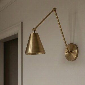 May include: A wall-mounted lamp with a brass finish, featuring a circular base plate, an adjustable two-part articulated arm, and a conical shade. The fixture is constructed from metal and is positioned to extend outward from the wall surface.