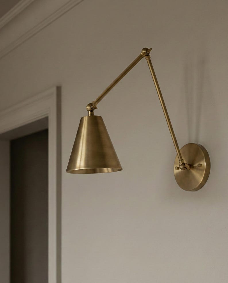 May include: A wall-mounted lamp with a brass finish, featuring a circular base plate, an adjustable two-part articulated arm, and a conical shade. The fixture is constructed from metal and is positioned to extend outward from the wall surface.