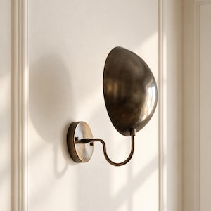 May include: A wall-mounted sconce featuring a dark bronze-toned, semi-spherical metal shade attached to a curved arm and a circular wall plate. The fixture has a smooth, metallic finish and is positioned to cast a soft shadow on the wall.