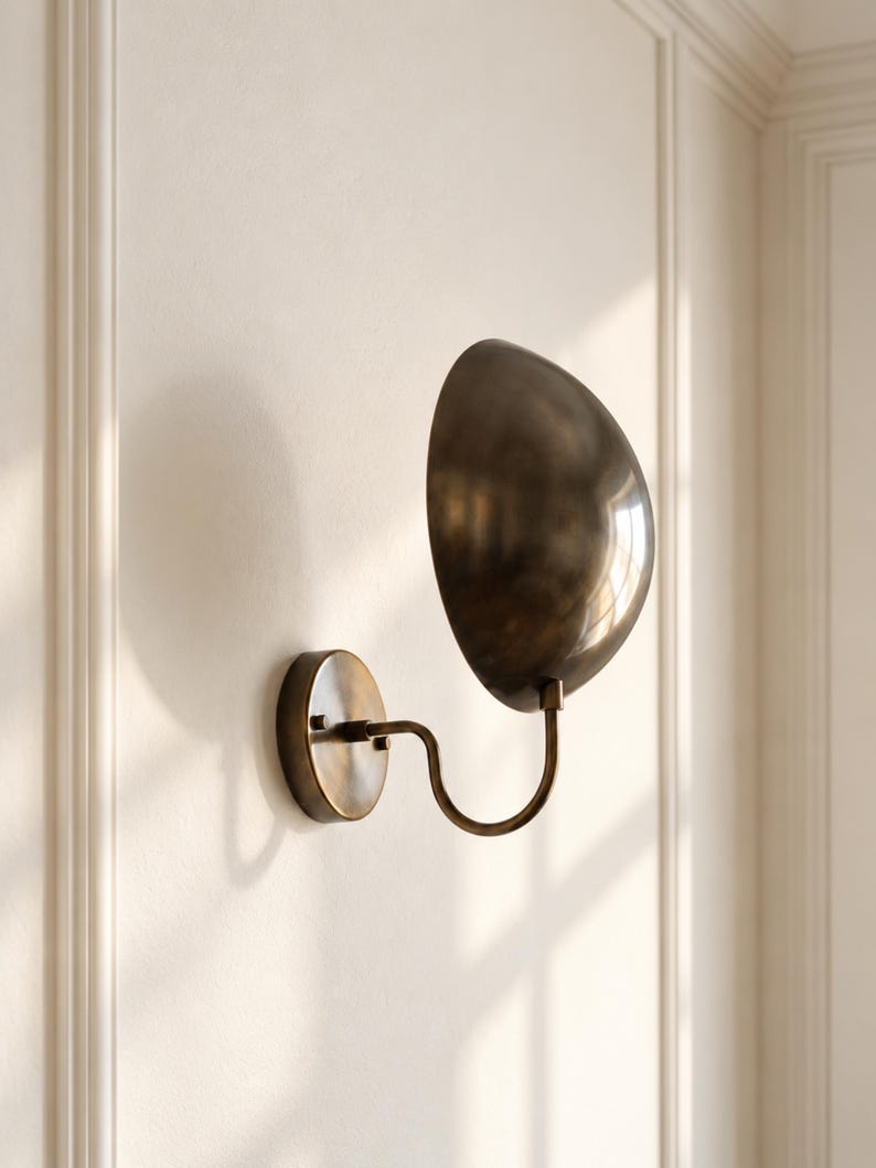 May include: A wall-mounted sconce featuring a dark bronze-toned, semi-spherical metal shade attached to a curved arm and a circular wall plate. The fixture has a smooth, metallic finish and is positioned to cast a soft shadow on the wall.