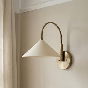 May include: A wall-mounted sconce featuring a conical off-white shade and a curved brass arm. The sconce is fixed to a round brass base. This indoor lighting fixture provides ambient illumination.