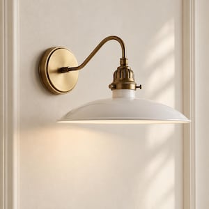 May include: A wall-mounted light fixture featuring a circular brass backplate, a curved brass arm, and a wide, shallow white enamel shade. The socket housing is finished in a matching brass tone, connecting the arm to the white metal shade.