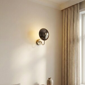 May include: A wall-mounted light fixture featuring a circular, dark metallic shade attached to a curved brass-toned arm and a round base plate. The lamp emits a warm, soft glow against the wall, highlighting its polished finish.