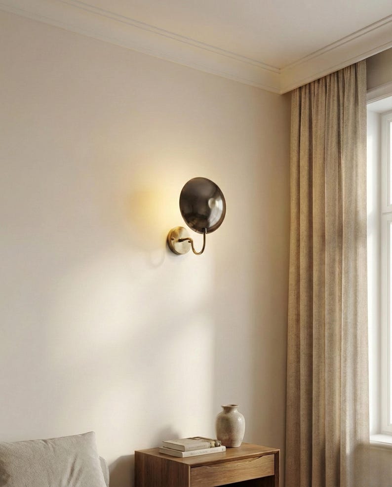May include: A wall-mounted light fixture featuring a circular, dark metallic shade attached to a curved brass-toned arm and a round base plate. The lamp emits a warm, soft glow against the wall, highlighting its polished finish.