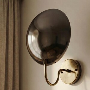 May include: A wall-mounted lamp featuring a large, circular, dark metallic reflective shade attached to a curved, antique brass-toned arm that connects to a matching circular base plate mounted flush against the wall surface.