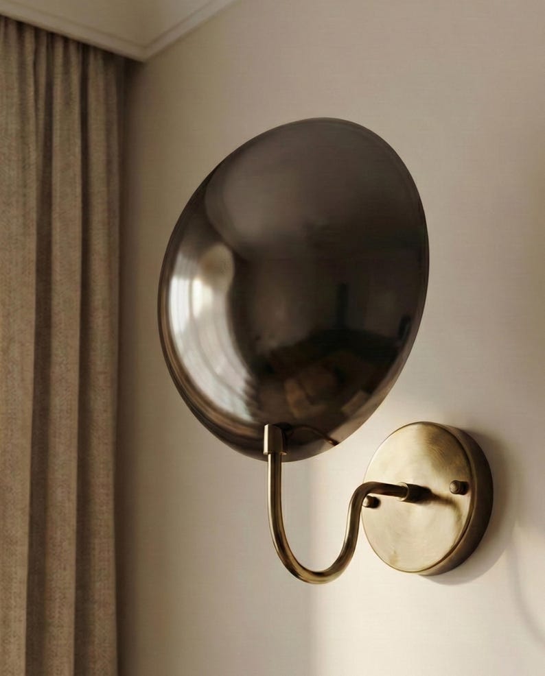 May include: A wall-mounted lamp featuring a large, circular, dark metallic reflective shade attached to a curved, antique brass-toned arm that connects to a matching circular base plate mounted flush against the wall surface.