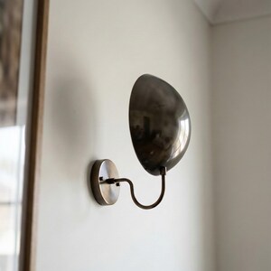 May include: A wall-mounted lamp featuring a circular metallic base and a curved, S-shaped arm that supports a large, polished, dark metallic dome-shaped shade. The fixture has a reflective finish and is attached to a plain, light-colored wall.
