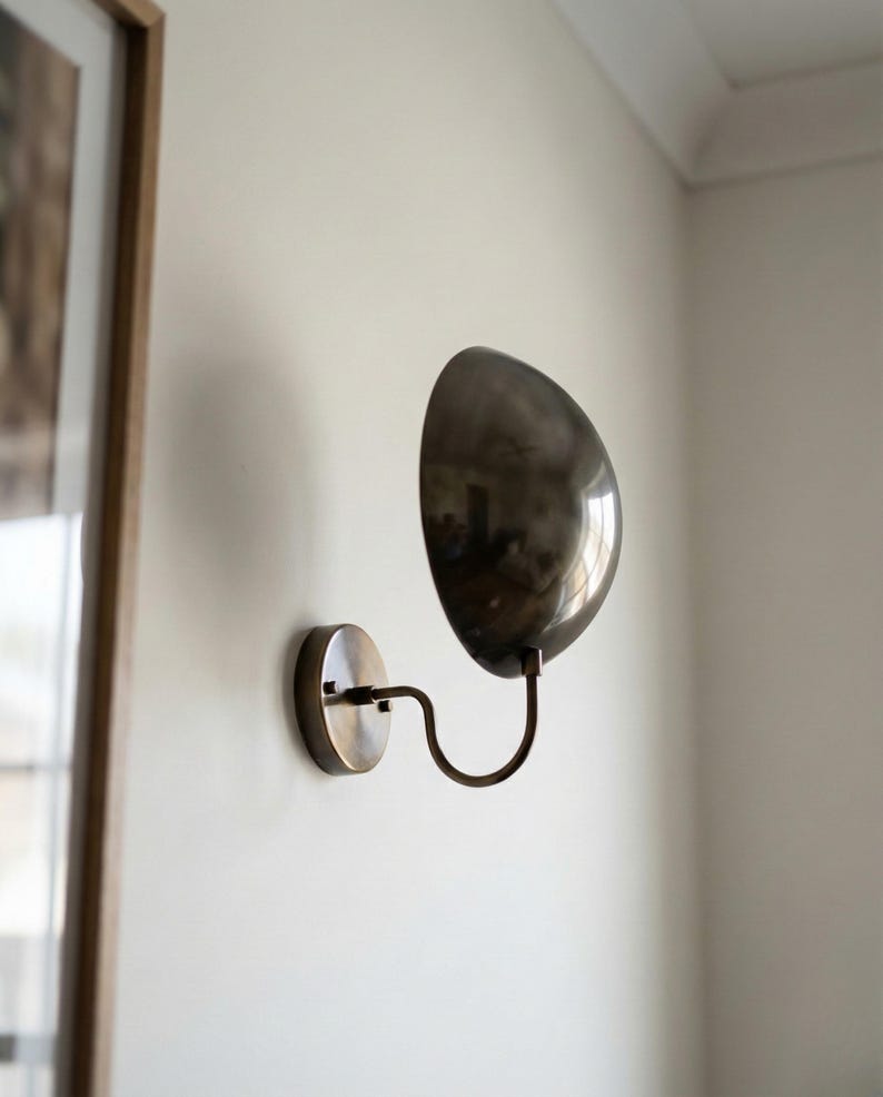 May include: A wall-mounted lamp featuring a circular metallic base and a curved, S-shaped arm that supports a large, polished, dark metallic dome-shaped shade. The fixture has a reflective finish and is attached to a plain, light-colored wall.
