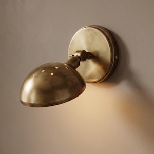 May include: A wall-mounted lamp with an antique brass finish, featuring a circular base and an adjustable, dome-shaped shade. The top of the shade has small, perforated holes that allow light to pass through, creating a soft, focused illumination.