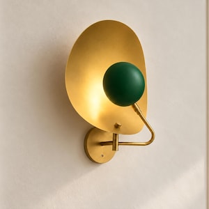May include: A wall sconce featuring a large, circular, brushed gold metal reflector plate. A dark green spherical glass shade is mounted on a curved gold arm that extends from a circular gold base plate attached to the wall.