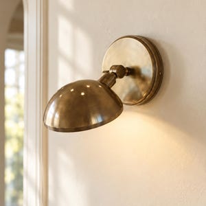 May include: A wall-mounted brass sconce featuring a circular backplate and a dome-shaped shade. The fixture includes an adjustable swivel joint that allows the light to be angled downward, casting a warm glow against the wall surface.