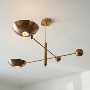 May include: A brass-toned ceiling light fixture with three bowl-shaped shades and three spherical accents. The fixture is suspended from the ceiling with a central vertical rod and horizontal arms. The light is on.