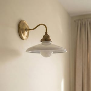 May include: A wall-mounted light fixture with a brass finish and a white, bowl-shaped shade. The fixture has a curved arm and a round base, mounted on a beige wall. The light bulb is visible inside the shade.