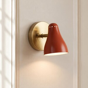 May include: A wall-mounted sconce featuring a matte burnt orange bell-shaped shade with a row of small circular perforations near the top, attached to a circular brushed brass backplate by a short horizontal arm.