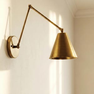 May include: A wall-mounted swing arm lamp with a brushed brass finish. The fixture features a circular wall plate, a long adjustable metal arm with a central hinge, and a conical shade that directs light downward from the end of the arm.