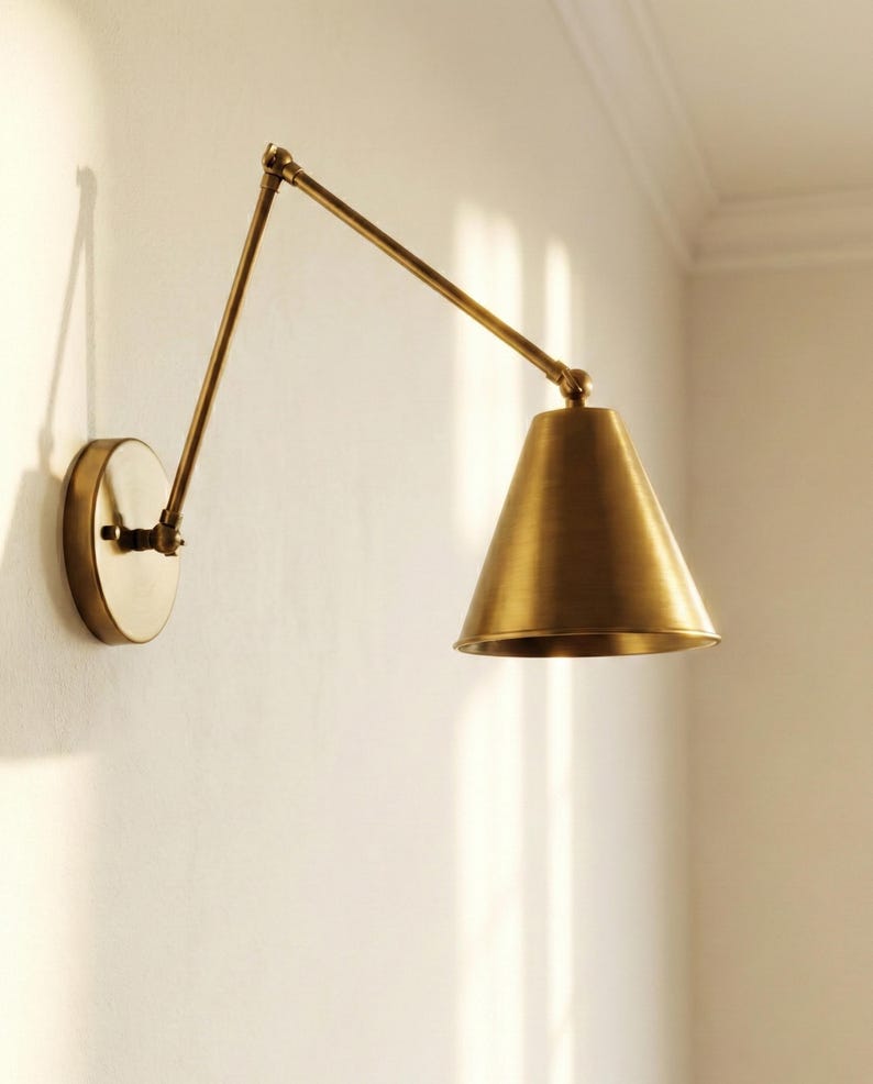 May include: A wall-mounted swing arm lamp with a brushed brass finish. The fixture features a circular wall plate, a long adjustable metal arm with a central hinge, and a conical shade that directs light downward from the end of the arm.