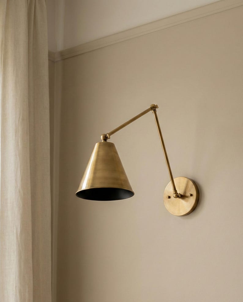 May include: A wall-mounted lamp featuring a brushed brass finish with a conical shade. The fixture includes an adjustable swing arm connected to a circular wall plate, with the interior of the cone shade finished in a contrasting matte black colour.